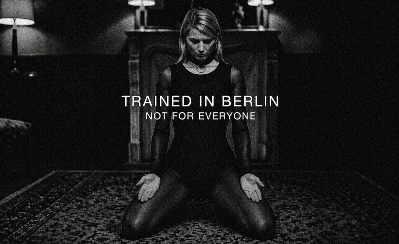 Submissive BDSM escort jO (Johanna Koltai) in Nadu position, kneeling with lowered gaze, calm and controlled in a dark, softly lit room – trained in Berlin