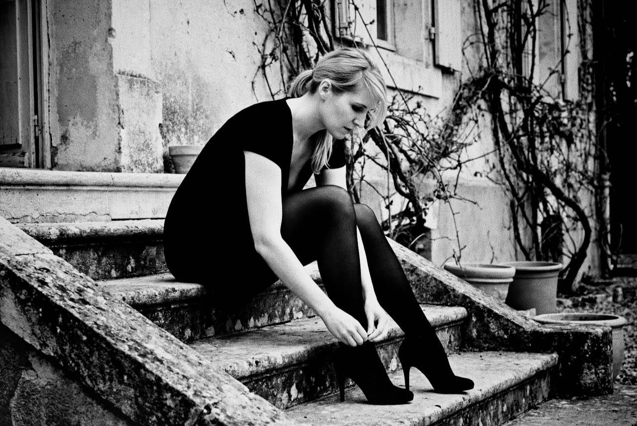 Submissive BDSM escort jO (Johanna Koltai) sitting on stone steps adjusting her heels in a private outdoor setting