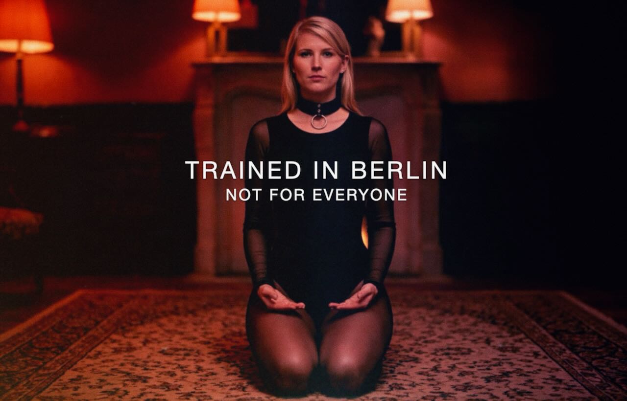 Submissive BDSM escort jO (Johanna Koltai) kneeling in a dark room with warm lighting, trained in Berlin, not for everyone