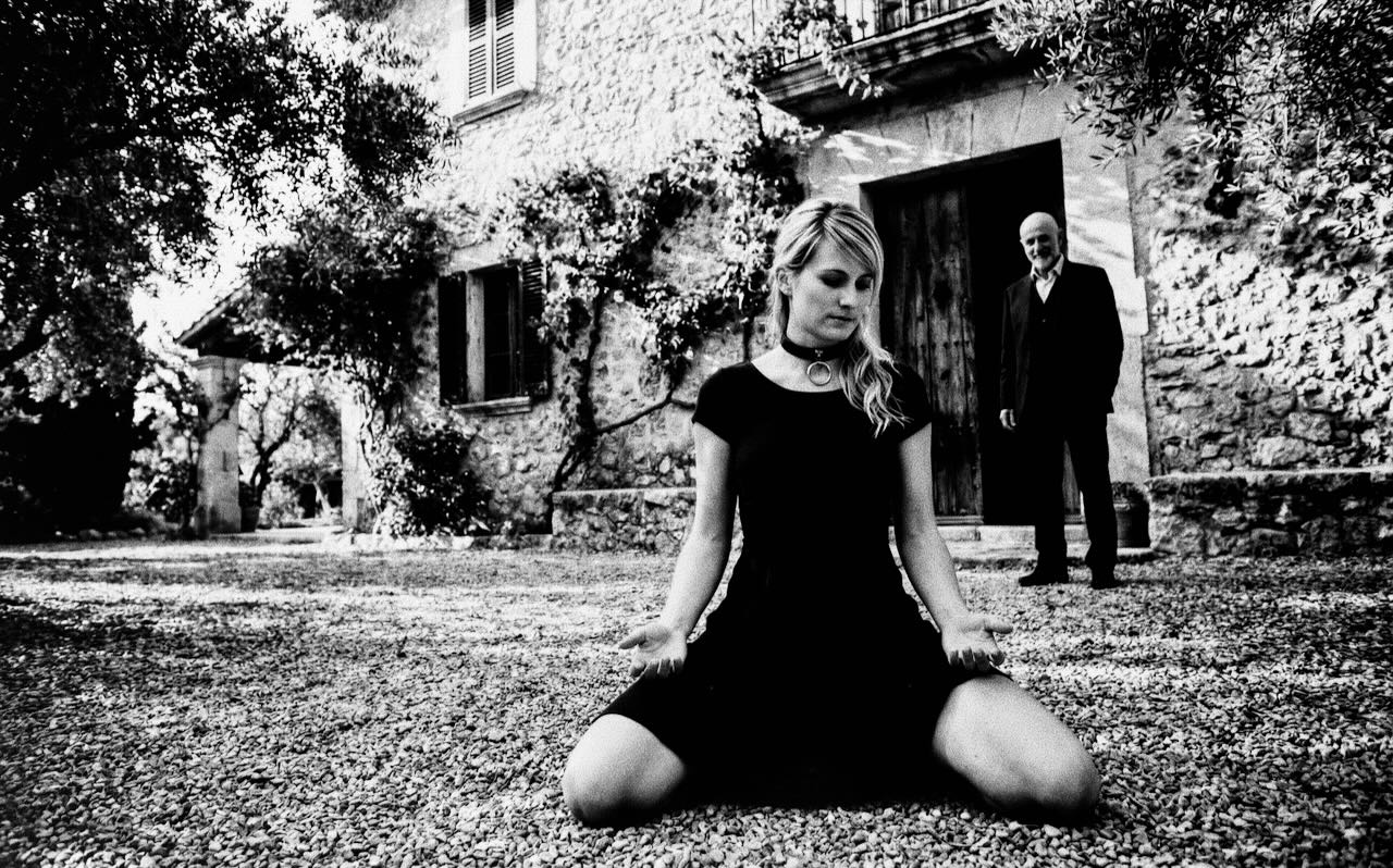 Submissive BDSM escort jO (Johanna Koltai) kneeling in front of a villa while a man stands in the background
