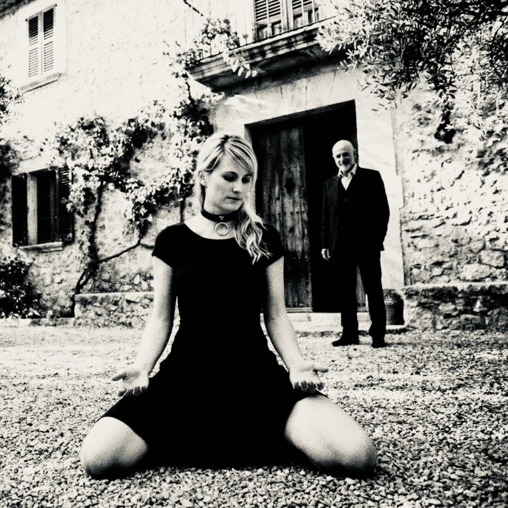 Escort Sklavin Zürich kniet in schwarzem Kleid ruhig auf Kies vor einem alten Haus, im Hintergrund steht ein Mann in der Tür.