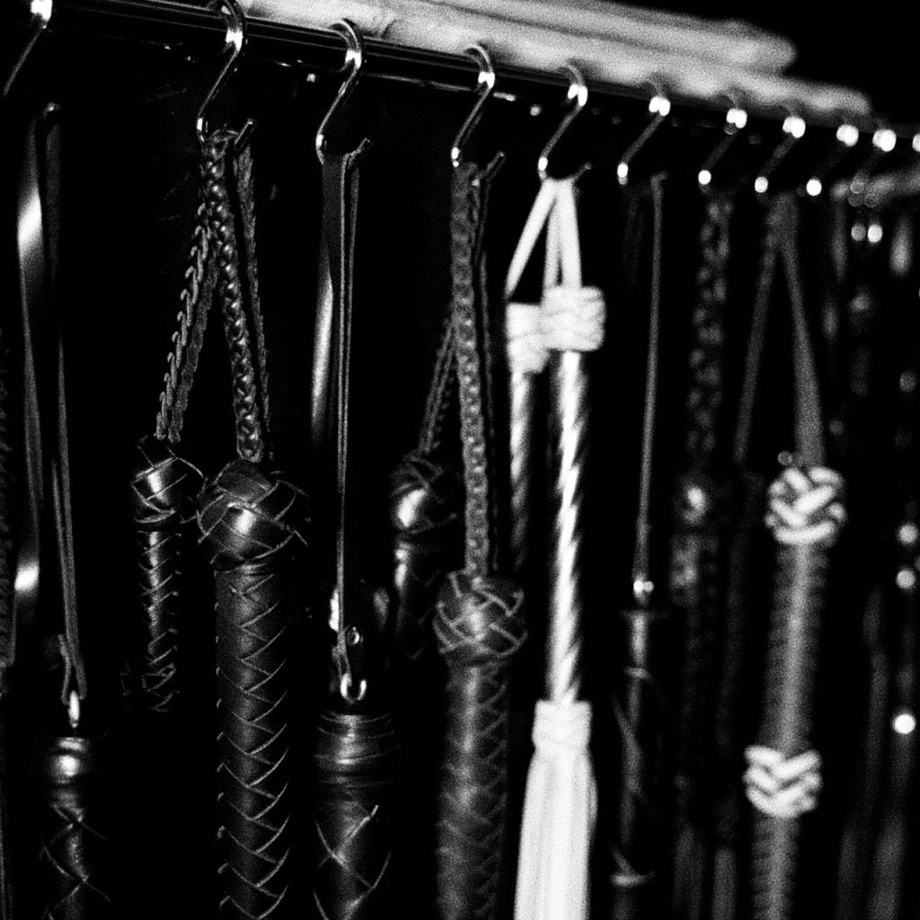 Black and white image of BDSM tools including whips and floggers hanging on a metal rack