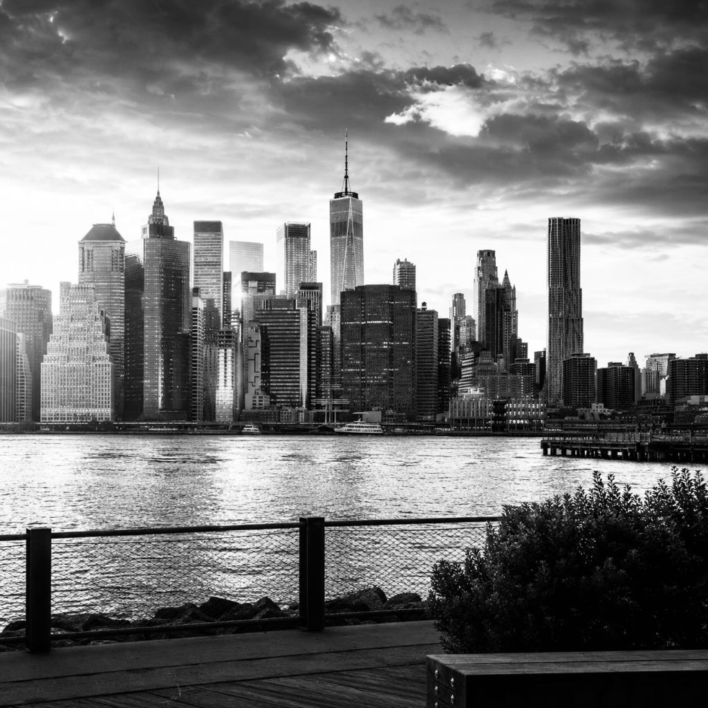 Manhattan skyline at sunset viewed from Brooklyn Bridge Park, with One World Trade Center illuminated by golden evening light and reflections shimmering across the East River.