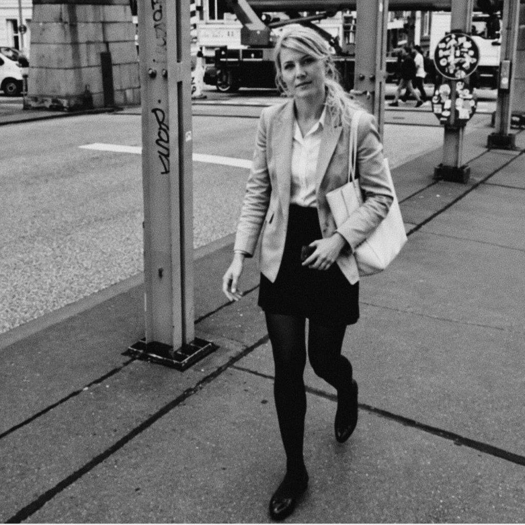 BDSM escort jO walking through the city in a blazer, short black skirt, and black tights.